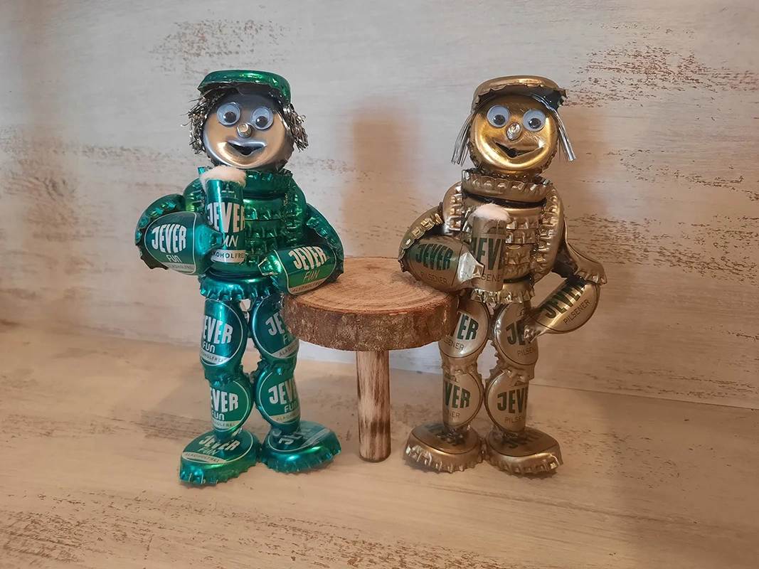 Zwei Figuren aus grünen und goldenen Kronkorken gefertigt, stehen neben einem kleinen Holztisch.