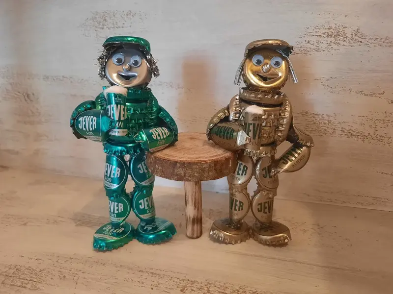 Zwei Figuren aus grünen und goldenen Kronkorken gefertigt, stehen neben einem kleinen Holztisch.