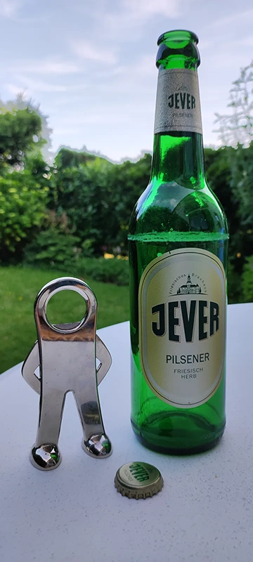 Jever Bierflasche steht neben einem metallenen Flaschenöffner in Figurenform. Im Hintergrund grüne Hecke.