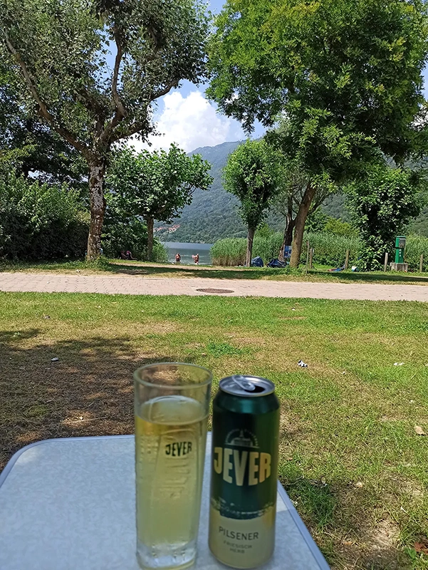 Ein Jever Glas und eine Dose Jever Pilsener stehen auf einem Tisch. Im Hintergrund Bäume und ein See mit Bergen.