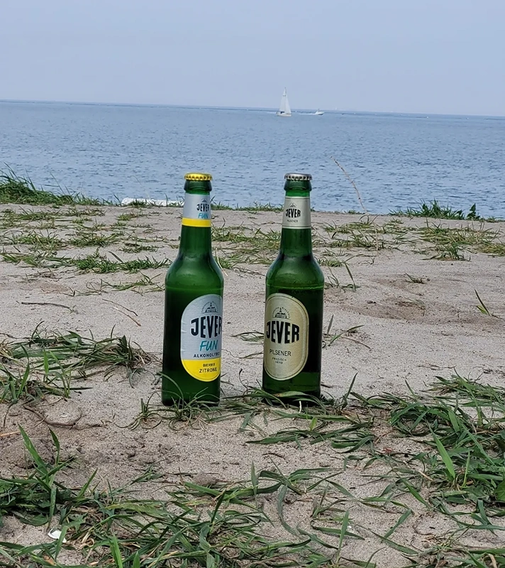 Zwei Jever Bierflaschen stehen auf sandigem Boden mit Gras. Im Hintergrund ist das Meer mit einem Segelboot zu sehen.