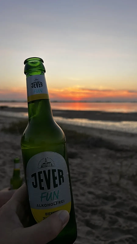 Eine Flasche Jever Fun alkoholfrei wird in einer Hand gehalten. Im Hintergrund Sonnenuntergang am Sandstrand.