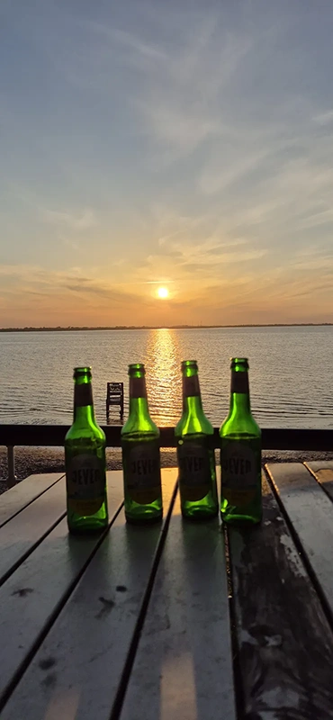 Vier Jever Flaschen stehen auf einem Holztisch. Im Hintergrund ist ein ruhiger See mit Sonnenuntergang.