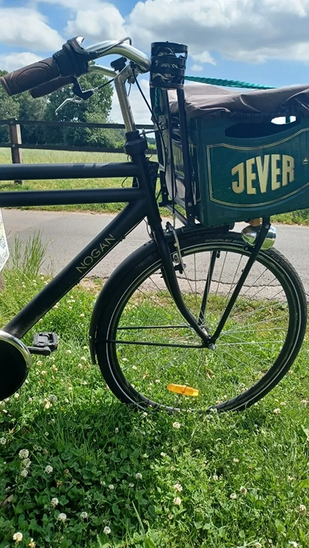 Ein Lastenfahrrad mit Jever Getränkekiste auf der Vorderseite.  Im Hintergrund eine Wiese bei leicht bewölktem Himmel.
