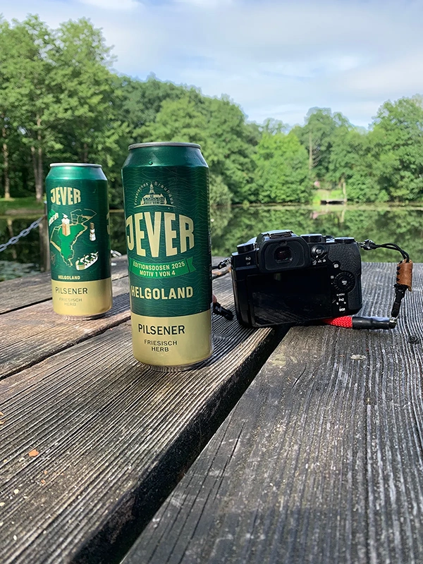 Zwei Jever Pilsener Dosen und eine Kamera stehen auf einem Holztisch. Im Hintergrund ist ein See mit Bäumen.