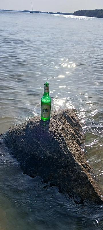 Eine Jever Flasche steht auf einem nassen Felsen im Wasser. Im Hintergrund glitzert die Wasseroberfläche in der Sonne.