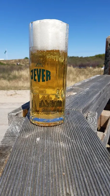 Ein Jever Glas mit Schaumkrone ist mit hellem Bier gefüllt. Im Hintergrund stehen Dünen und blauer Himmel.
