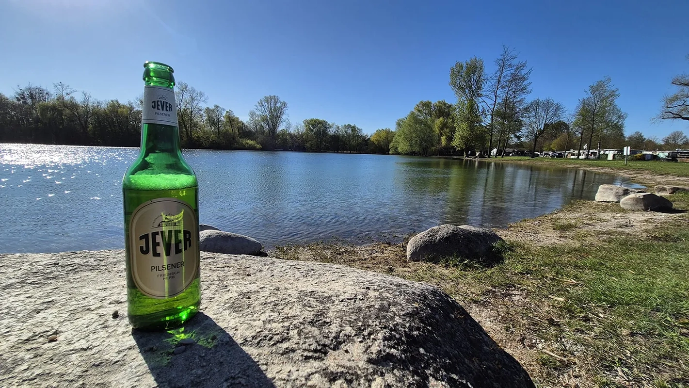 Jever Bierflasche steht auf einem großen Stein am Ufer eines ruhigen Sees. Im Hintergrund Bäume und ein Campingplatz.