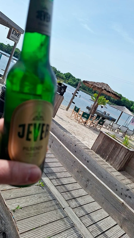 Jever Bierflasche im Vordergrund. Im Hintergrund Sandstrand mit Liegestühlen und See mit Palmen.