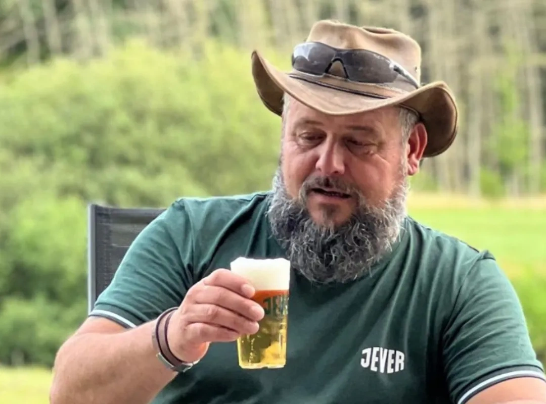 Mann mit Bart und Hut hält ein Jever in der Hand. Im Hintergrund sind Bäume und Wiese unscharf erkennbar.