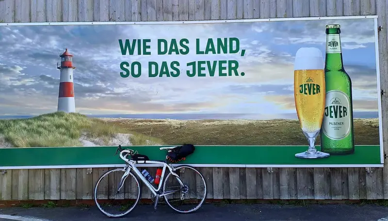 Ein weißes Fahrrad steht vor einem Jever Werbeplakat mit Jever Bierflasche und Jever Glas vor Dünen und Leuchtturm.