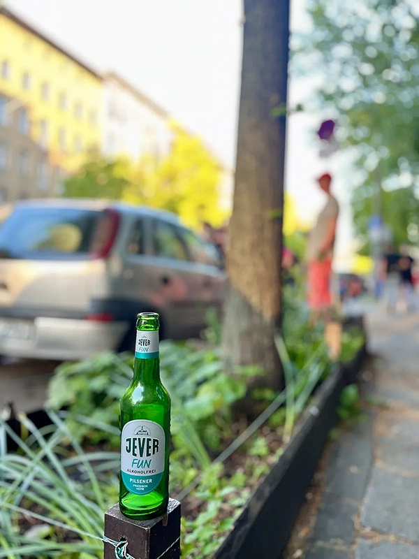 Jever Fun Flasche auf einem Holzpfosten im Vordergrund. Unscharfer Gehweg mit Auto und Bäumen im Hintergrund.