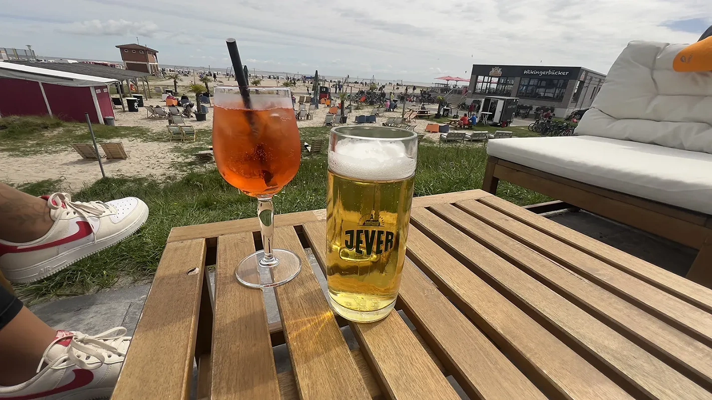 Ein Glas Jever Bier und ein Glas Spritz stehen auf einem Holztisch. Im Hintergrund ein belebter Strand mit Gastronomie.