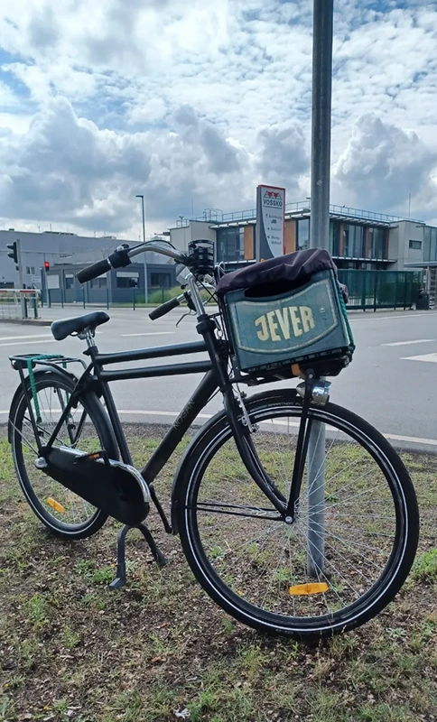 Schwarzes Herrenrad mit Jever Kasten am Lenker lehnt an einem Verkehrsschild. Im Hintergrund moderne Firmengebäude.