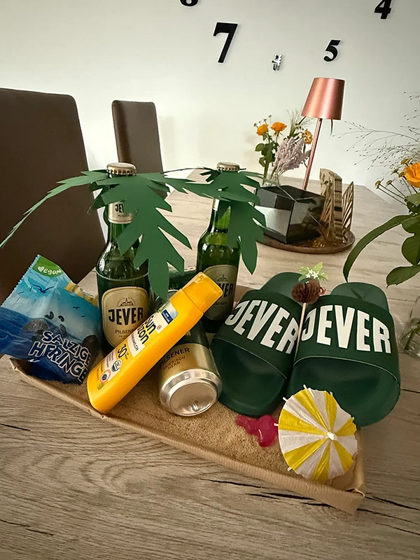 Drei Bierflaschen, Badeschlappen, Sonnencreme und Süßigkeiten liegen auf einem Tablett mit Dekosand.