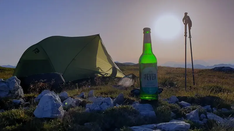 Grünes Zelt, eine Jever Flasche und zwei Wanderstöcke auf einer Wiese. Im Hintergrund tiefstehende Sonne und Berge.