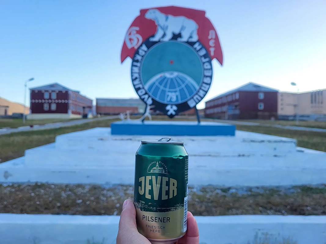 Eine Hand hält eine Jever Pilsener Dose vor einem Denkmal mit Eisbären Emblem. Im Hintergrund sind rote Kasernengebäude.