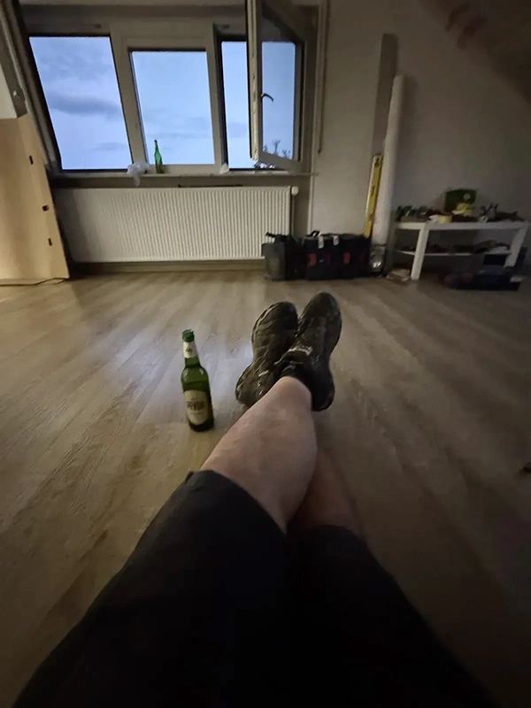 Person mit überschlagenen Beinen sitzt auf Holzboden, daneben eine Jever Flasche und Werkzeug vor geöffnetem Fenster.