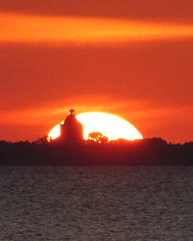Eine große untergehende Sonne steht hinter einem Kirchturm mit umliegenden Bäumen. Im Vordergrund ist eine Wasserfläche.