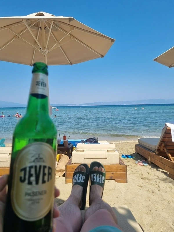 Jever Flasche vor ausgestreckten Beinen auf einer Liege am Sandstrand. Im Hintergrund Meer und Sonnenschirme.