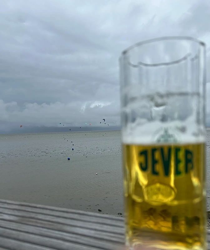 Ein halbvolles Jever Glas steht auf einem hölzernen Tisch. Im Hintergrund bedeckter Himmel und ein Meer mit Kitesurfern.