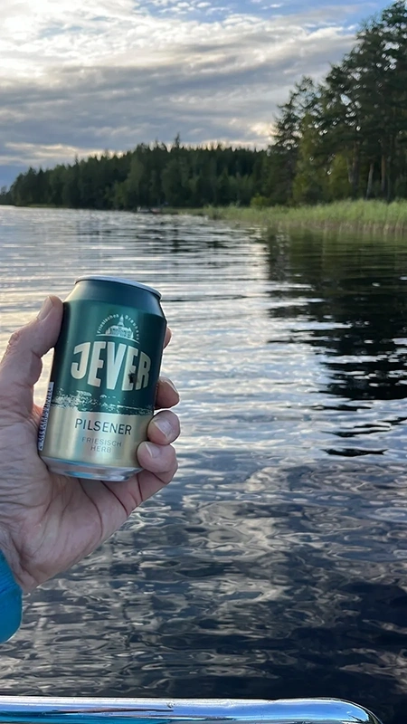 Eine Hand hält eine Jever Pilsener Dose über einer Wasserfläche. Im Hintergrund dichter Wald unter wolkigem Himmel.