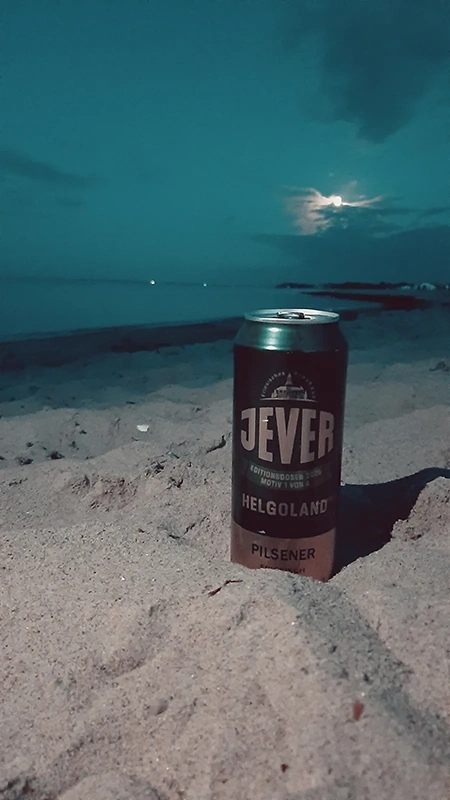 Eine geöffnete Bierdose steht auf hellem Sand. Im Hintergrund ist das Meer mit Mondschein und Wolken zu sehen.