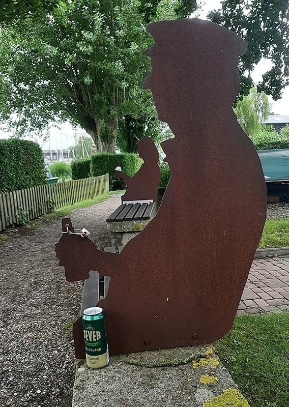 Metallskulptur eines sitzenden Mannes mit Flasche auf einer Bank. Im Hintergrund Bäume und weitere Silhouetten.