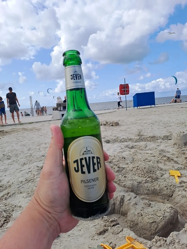 Jever Pilsener Flasche wird in einer Hand am Strand gehalten. Im Hintergrund Strandbesucher und das Meer.