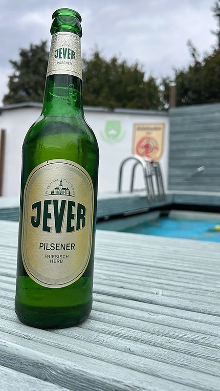 Jever Pilsener Flasche auf grauer Holzfläche. Im Hintergrund ein Pool mit Leiter und eine weiße Wand mit Logos.