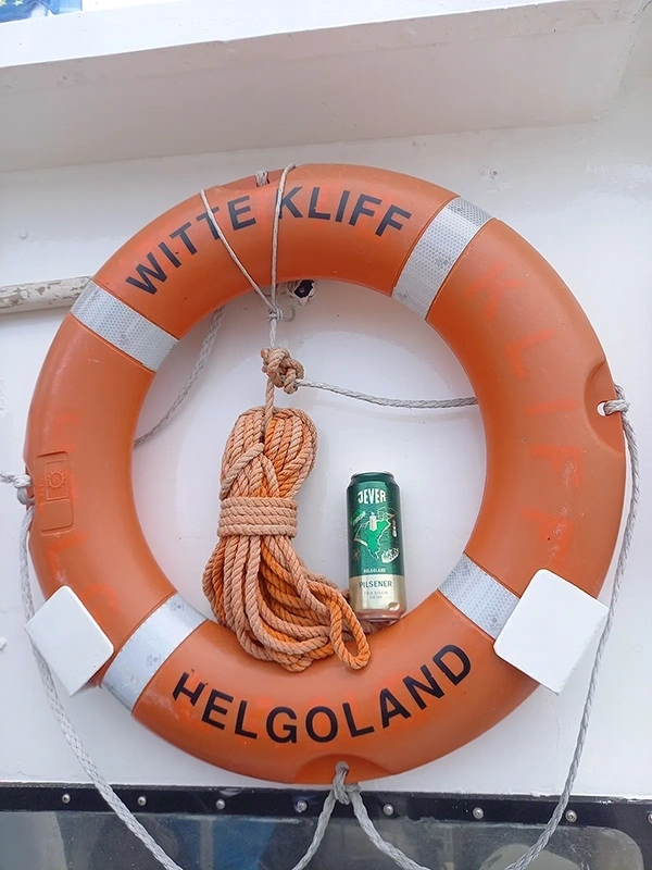 Oranger Rettungsring mit schwarzer Helgoland Aufschrift und Seil, in der Mitte eine Jever Bierdose.