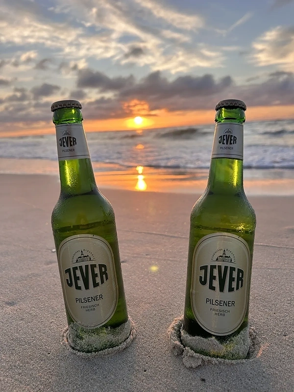 Zwei Jever Flaschen stecken im Sand. Im Hintergrund Sonnenuntergang über dem Meer mit Wolkenhimmel.