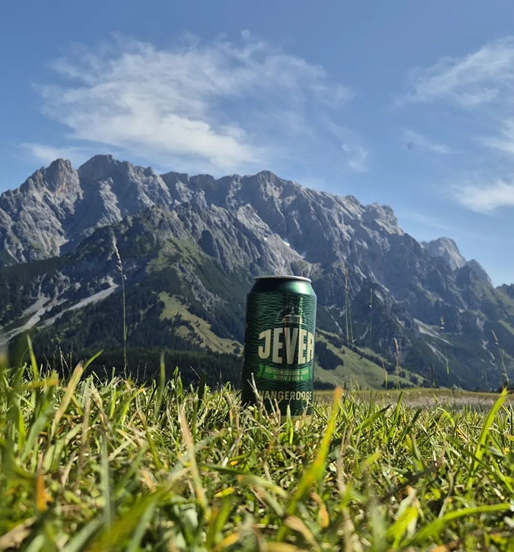 Jever Pilsener Dose steht auf einer Wiese. Im Hintergrund hohe felsige Berge unter blauem Himmel.
