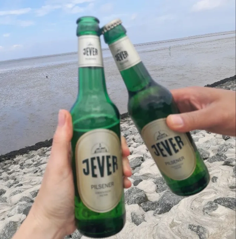 Zwei Hände stoßen mit  Jever Pilsener Flaschen an. Im Hintergrund ein steiniger Strand und das Wattenmeer.