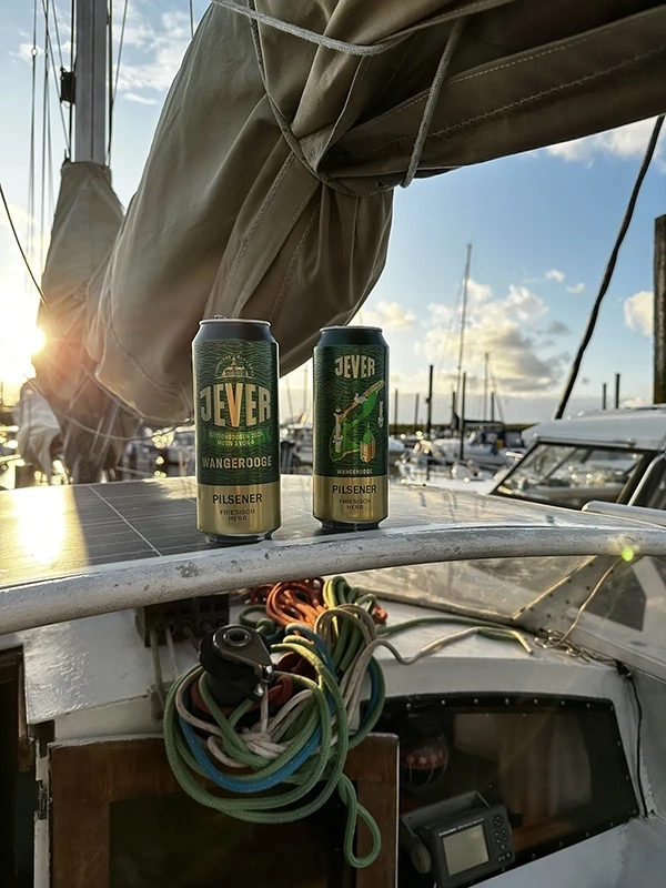 Zwei Jever Pilsener Dosen stehen auf einem Boot vor aufgewickelten Seilen. Im Hintergrund liegen Segelboote im Hafen.