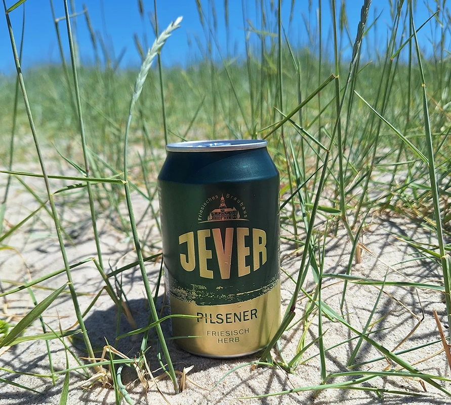 Eine Jever Pilsener Dose steht auf sandigem Boden zwischen dünnen Grashalmen. Im Hintergrund blauer Himmel.