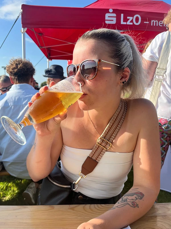 Junge Frau mit Sonnenbrille trinkt aus einem Jever Bierglas. Im Hintergrund Menschen und ein rotes Zelt mit Aufschrift.