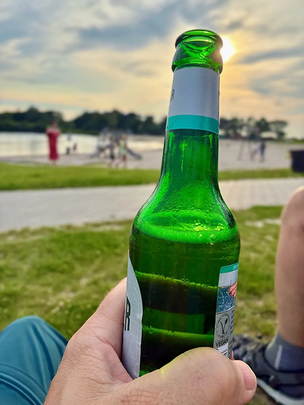 Eine Hand hält eine Jever Flasche. Im Hintergrund liegt ein Sandstrand mit See, Bäumen und spielenden Menschen.