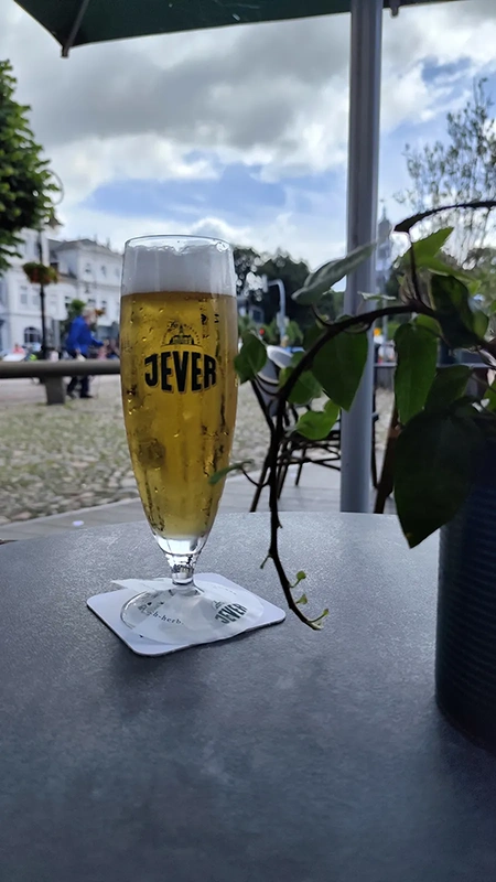 Ein Jever Glas steht auf einem Tisch, daneben eine Pflanze. Im Hintergrund ist ein gepflasterter Platz mit Personen.