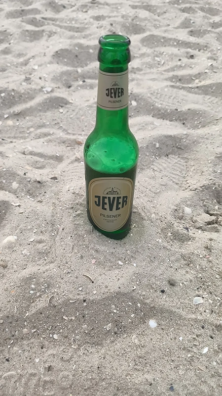 Jever Pilsener Flasche steht aufrecht im Sand. Der Hintergrund besteht aus feinem Sand mit Muschelresten.