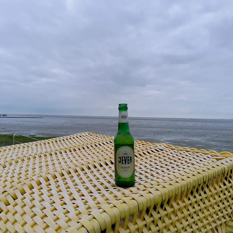 Jever Pilsener Flasche steht auf gelb geflochtenem Strandkorb. Im Hintergrund Wattlandschaft unter bewölktem Himmel.