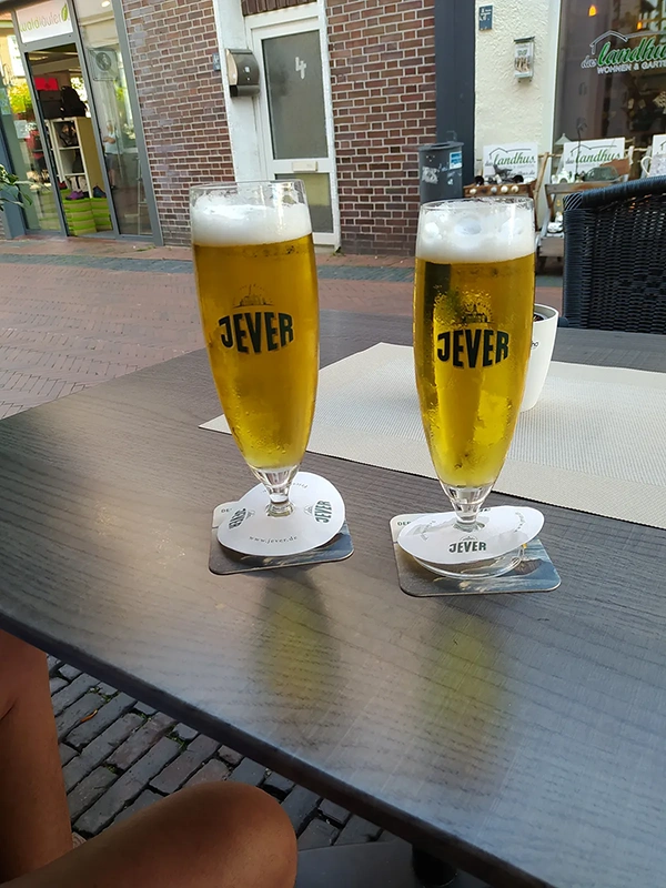 Zwei gefüllte Jever Gläser stehen auf einem Holztisch. Im Hintergrund eine Einkaufsstraße mit Läden.