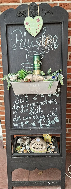 Eine schwarze Staffelei mit Kreidetafel, bepflanztem Zinkkasten und Jever Dose als Dekoration vor roter Ziegelwand.