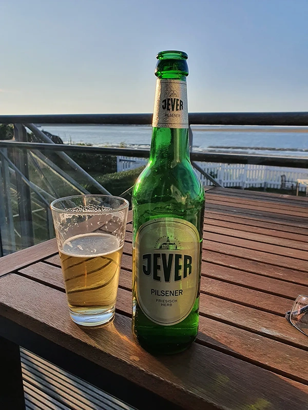 Jever Flasche und gefülltes Bierglas auf einem Holztisch. Im Hintergrund Meerblick mit Geländer und weißem Zaun.