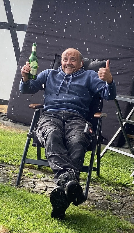 Ein Mann sitzt im Regen auf einem Campingstuhl, hält eine Jever Flasche und zeigt den Daumen hoch.