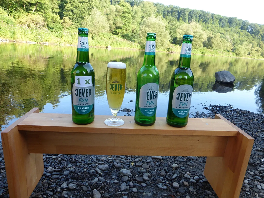 Drei Flaschen alkoholfreies Jever Pilsener auf einer Holzbank, mittig ein gefülltes Bierglas vor einer Flusslandschaft.