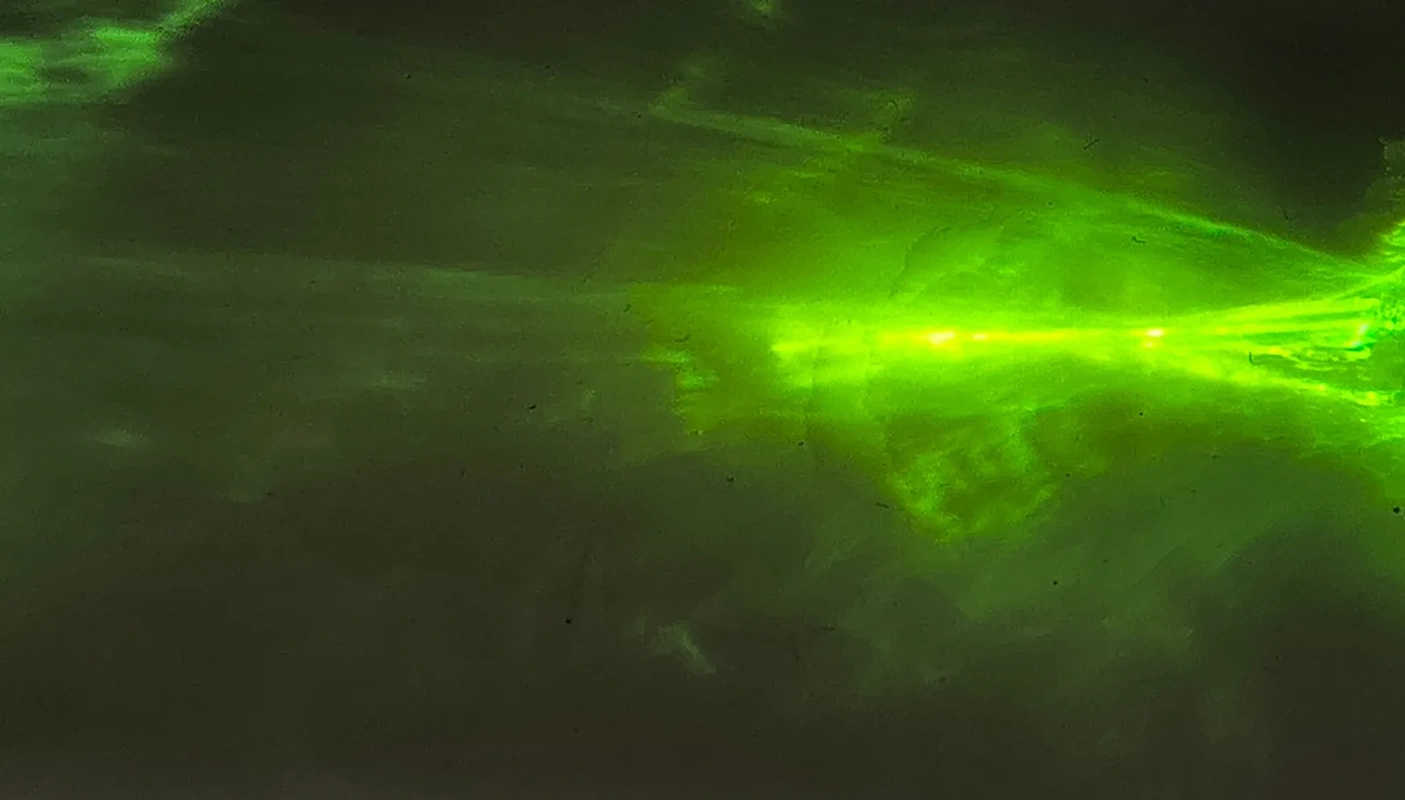 Grüner Lichtstrahl mit leuchtendem Zentrum auf dunkler Fläche. Im Hintergrund diffuse Strukturen.