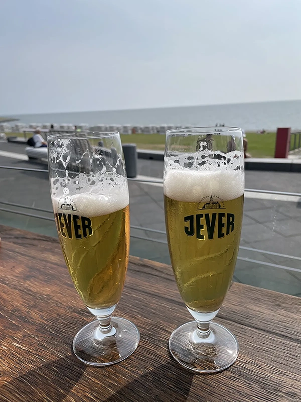 Zwei gefüllte Jever Gläser stehen auf einem Holztisch. Im Hintergrund befinden sich Rasenfläche und Strandkörbe am Meer.