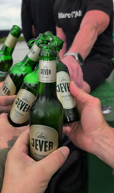 Sechs Jever Flaschen werden in einer Gruppe angestoßen. Im Hintergrund sind Arme und Oberkörper von Personen.