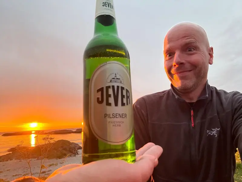 Eine Jever Pilsener Flasche wird in der Hand gehalten. Im Hintergrund Sonnenuntergang am Meer mit Felsen und Strand.